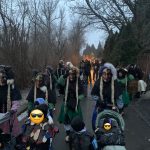 Fasnetsfiguren und Kinder in traditionellen Gewändern und Masken ziehen in der Dämmerung einen winterlichen Weg entlang.
