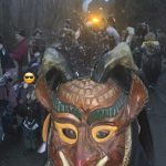 Große Hornmaske einer Fasnetsfigur im Vordergrund, dahinter eine Gruppe von Teilnehmern auf einem winterlichen Weg bei Abendbeleuchtung.