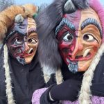 Fasnetsfiguren in traditionellen Gewändern mit Zöpfen und farbigen Häsern gehen einen winterlichen Weg entlang.