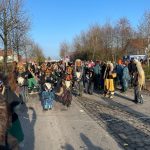 Fasnetsfiguren in traditionellen Masken und farbigen Häsern laufen bei sonnigem Wetter eine breite Straße entlang, umgeben von vielen Zuschauern.