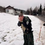 Fasnetsteilnehmer in traditionellem Häs steht im Schnee und erfrischt sich am Wegesrand in winterlicher Dorfumgebung.