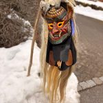 Traditionelle geschnitzte Fasnetsmaske mit Hörnern und langen Haaren steht an einem Stab im Schnee vor einem Gehweg.