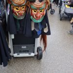 Zwei traditionelle Fasnetsmasken mit Hörnern und Stoffplättchen hängen an der Vorderseite eines Transportwagens während eines Umzugs.