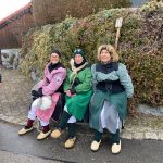 Drei Fasnetsfiguren in traditionellen Gewändern sitzen auf einer Bank am Straßenrand neben einem Schild mit der Nummer 25.