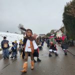 Mehrere Fasnetsgruppen in bunten traditionellen Gewändern versammeln sich auf einer winterlichen Dorfstraße während eines Umzugs.