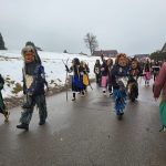 Mehrere Fasnetsfiguren in traditionellen Masken und Plättlehäs laufen während eines Umzugs eine winterliche Landstraße entlang.