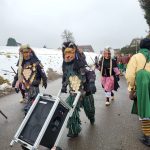 Fasnetsfiguren in traditionellen Masken und Plättlehäs laufen über eine winterliche Landstraße, eine Figur zieht einen mobilen Lautsprecherwagen.
