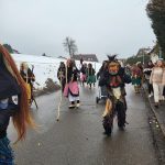 Fasnetsfiguren in traditionellen Masken und Häsern laufen während eines Winterumzugs eine Dorfstraße entlang.
