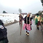 Fasnetsfiguren in traditionellen Masken und Gewändern laufen während eines Umzugs eine winterliche Dorfstraße entlang.