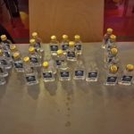 Mehrere Gruppen kleiner Getränkeflaschen mit gelben Verschlüssen stehen geordnet auf einem Tisch.