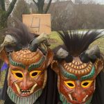 Zwei traditionelle Fasnetsmasken mit Hörnern und farbigen Plättchen stehen vor einem Schild mit der Nummer 4 in einer winterlichen Landschaft.