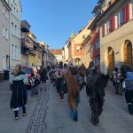 Fasnetsgruppe in traditionellen Häsern marschiert durch eine enge Altstadtstraße mit Häusern und Sonnenschein.