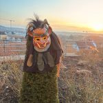 Traditionelle Fasnetsmaske mit Stoff- und Fellbesatz auf einem Busch, im Hintergrund Landschaft und Sonnenuntergang.