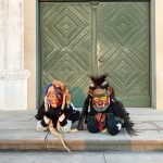 Zwei Fasnetsfiguren in traditionellen Masken und Häsern sitzen auf Stufen vor einer großen grünen Holztür mit Rundbogen.