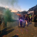 Fasnetsteilnehmer in traditionellen Masken ziehen am Abend durch eine Straße, begleitet von Rauch- und Lichteffekten sowie zahlreichen Zuschauern.