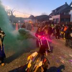 Fasnetsteilnehmer in traditionellen Masken ziehen am Abend durch eine Straße, begleitet von Rauch- und Lichteffekten sowie zahlreichen Zuschauern.