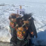 Drei bunt geschnitzte Fasnets-Holzmasken stehen auf einem Transportwagen vor einer verschneiten Winterlandschaft mit einem Schild der Nummer 29.