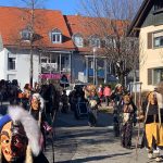 Fasnetsteilnehmer in geschnitzten Masken und traditionellen Häsern ziehen bei Sonnenschein durch eine Dorfstraße, weitere Figuren und Zuschauer im Hintergrund.
