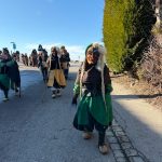 Hästräger in traditionellen Fasnetsgewändern laufen bei blauem Himmel durch eine Dorfstraße, weitere Teilnehmer folgen im Hintergrund
