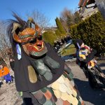 Fasnetsteilnehmer in traditionellen Holzmasken und farbigem Häs laufen bei sonnigem Wetter entlang einer Wohnstraße während eines Umzugs.