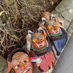 Mehrere bunt bemalte, traditionelle Fasnets-Holzmasken mit Fell- und Stoffelementen liegen am Rand eines Gehwegs zwischen Mauer und Sträuchern.