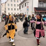 Traditionelle Fasnetsfiguren in farbigen Kostümen bewegen sich durch eine Altstadtstraße, begleitet von Zuschauenden und geschmückten Häusern.