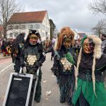 Mehrere traditionelle Fasnetsfiguren in schuppenartigen Kostümen ziehen bei einem Straßenumzug vorbei, begleitet von einem Lautsprecherwagen und zahlreichen Zuschauenden.