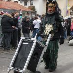 Traditionelle Fasnetsfigur mit schuppenartigem Häs zieht einen großen Lautsprecherwagen während eines Straßenumzugs, umgeben von Zuschauenden.