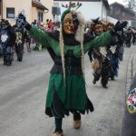Traditionelle Fasnetsfigur in grünem Häs mit ausgebreiteten Armen führt eine Gruppe maskierter Teilnehmender bei einem Straßenumzug an.