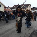 Fasnetsfigur in traditionellem Häs mit schuppenartigen Stoffplatten und geschnitzter Holzmaske schreitet bei einem Straßenumzug vorwärts.