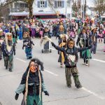 Traditionelle Fasnetsfiguren mit kunstvollen Masken und farbigen Häs treten auf einer Straße zwischen vielen Zuschauenden auf.