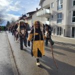 Traditionelle Fasnetsfiguren in farbigen Gewändern und mit Holzstäben laufen bei einem Straßenumzug durch ein modernes Wohngebiet.
