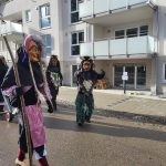 Fasnetsfiguren in traditionellen Masken und farbigen Gewändern laufen während eines Umzugs durch ein modernes Wohngebiet.