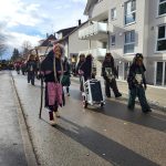 Traditionelle Fasnetsfiguren mit Masken und Kostümen laufen während eines Umzugs durch ein Wohngebiet und ziehen einen Lautsprecherwagen mit.